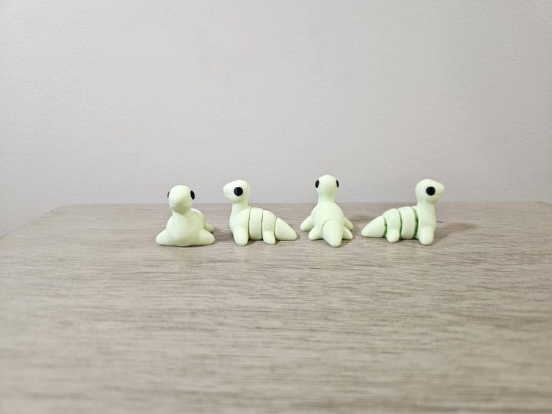 May include: Four small, light green clay dinosaur figurines with black eyes are arranged on a wooden surface. The dinosaurs are in various poses, some sitting and others standing. The figurines are handmade.