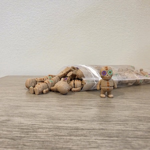 May include: A collection of small, light brown, humanoid figurines made of a textured material, each featuring a round head with two distinct, colorful circular eyes. One figurine stands upright in the foreground, while others are inside a clear plastic bag.