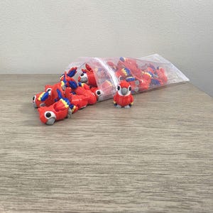 May include: A collection of small, red parrot figurines with blue and yellow accents, spilling from a clear plastic bag. One figurine stands upright, showcasing the details of the bird's design. The figurines are on a wooden surface.
