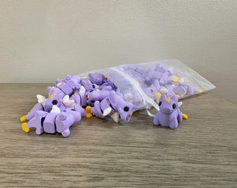 May include: A collection of small, purple dragon figurines with white wings and yellow accents. Some dragons are spilling out of a mesh bag, while others are scattered on a wooden surface. The dragons have black eyes.