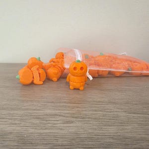 May include: A collection of small, orange, pumpkin-shaped figurines with green stems and carved facial features. Several of these miniature figures are scattered outside of a translucent, white drawstring pouch, while others remain inside the bag.