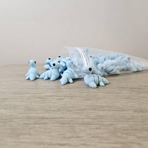 May include: Light blue polymer clay Nessie figurines with black eyes and segmented bodies are scattered on a wooden surface, with more inside a clear plastic bag. The figurines are a whimsical craft item.