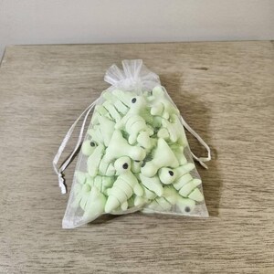 May include: A collection of light green dinosaur-shaped objects with black eyes, contained within a sheer white drawstring bag. The dinosaurs have a segmented body design. The bag is tied with a white ribbon.
