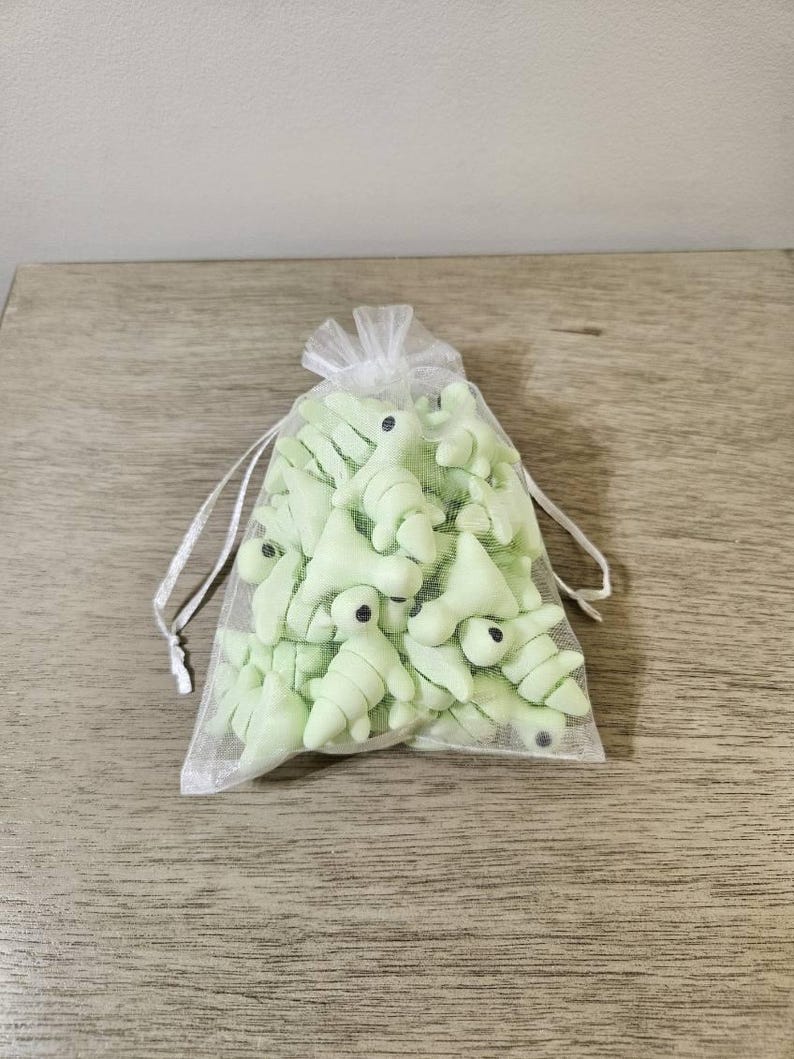 May include: A collection of light green dinosaur-shaped objects with black eyes, contained within a sheer white drawstring bag. The dinosaurs have a segmented body design. The bag is tied with a white ribbon.