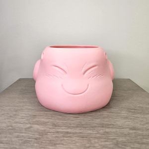 May include: A light pink planter shaped like a smiling face. The planter has a round opening and is made of a smooth material. The face has etched details for the eyes and mouth. The planter is sitting on a wooden surface.