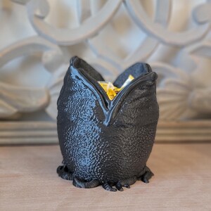 May include: A black, textured decorative container shaped like a creature's head, with a wide opening at the top. The container is filled with yellow and white items. The container is sitting on a wooden surface.