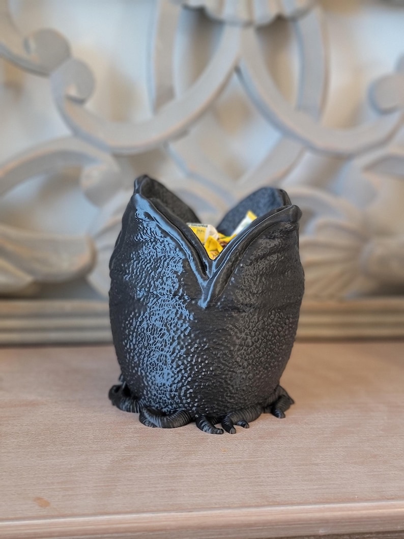 May include: A black, textured decorative container shaped like a creature's head, with a wide opening at the top. The container is filled with yellow and white items. The container is sitting on a wooden surface.