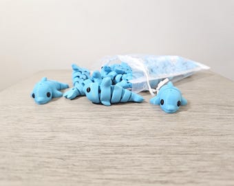May include: Light blue dolphin-shaped erasers scattered on a wooden surface, with some spilling from a clear bag. The erasers have black eyes and are a solid, pastel blue color. The bag contains more blue eraser pieces.