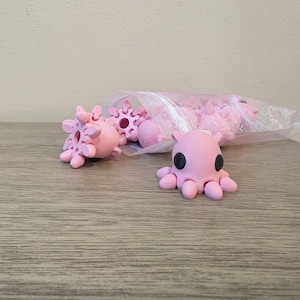 May include: A collection of small, light pink octopus figurines with black circular eyes. One figurine is positioned in the foreground, while several others are contained within a clear plastic bag behind it on a flat surface.