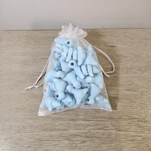 May include: A sheer, white drawstring bag filled with light blue plush shark toys. Each shark has black eyes and white accents. The bag is sitting on a light brown wooden surface.