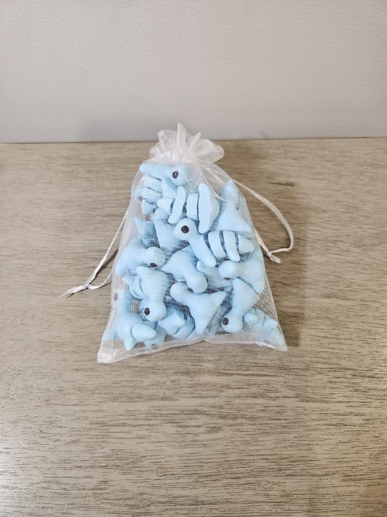May include: A sheer, white drawstring bag filled with light blue plush shark toys. Each shark has black eyes and white accents. The bag is sitting on a light brown wooden surface.
