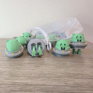 May include: A collection of green alien figurines with gray and yellow accents. The aliens are in a UFO-like design, some with legs. A few are inside a white mesh bag, suggesting they are sold as a set. The figurines are on a wooden surface.