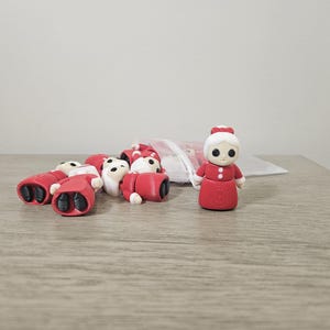 May include: A collection of red and white Christmas-themed erasers, including a standing Mrs. Claus figurine. The erasers are scattered on a wooden surface, with some spilling from a mesh bag. The Mrs. Claus figurine is red with white accents.