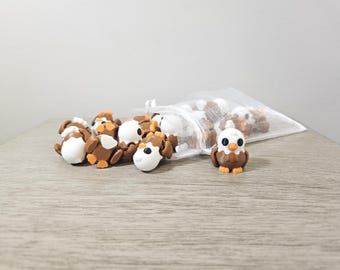 May include: A collection of small, decorative animal figurines. The figurines include brown and white dogs and a brown, white, and orange owl. Some figurines are in a clear mesh bag, while others are scattered on a wooden surface.