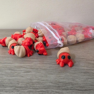 May include: A collection of small, three-dimensional printed hermit crab figurines, each featuring a bright red body with black eyes and a tan, textured spiral shell. Several crabs are clustered together, while others remain inside a clear bag.