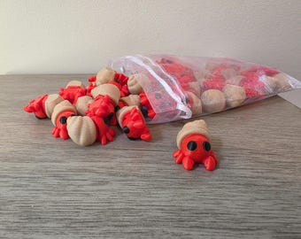 May include: A collection of small, three-dimensional printed hermit crab figurines, each featuring a bright red body with black eyes and a tan, textured spiral shell. Several crabs are clustered together, while others remain inside a clear bag.