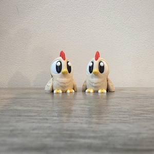 May include: Two small, beige-colored chicken figurines with large, expressive black and white eyes, a bright red comb on top of their heads, and a small yellow beak. Each chicken has visible textured details on its body and small yellow feet.
