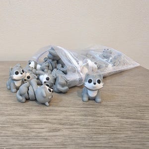 May include: A collection of small, gray and white wolf-shaped figurines with large black eyes, spilling out of a translucent white mesh drawstring bag. Each figurine features a seated posture with a white chest and face detail against a gray body.