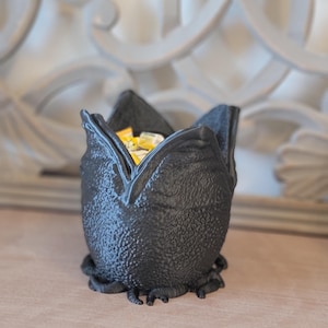 May include: A black, textured decorative bowl shaped like a flower. The bowl is filled with yellow and white items. The bowl has a unique, organic design and sits on a wooden surface.