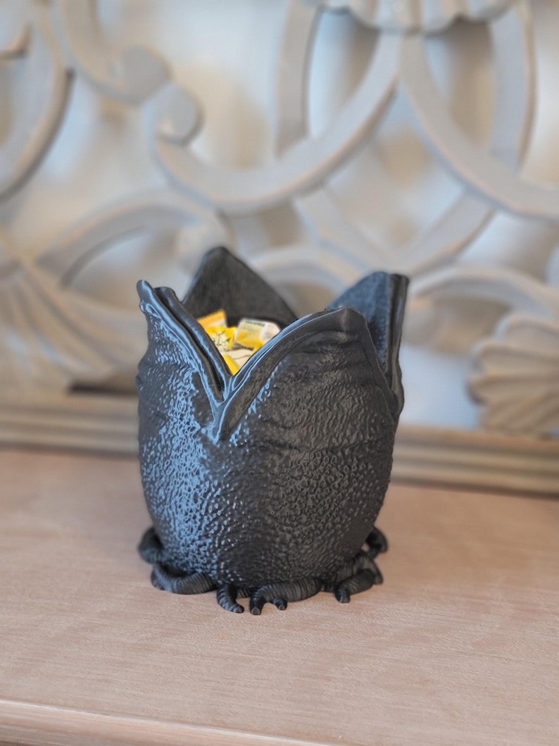 May include: A black, textured decorative bowl shaped like a flower. The bowl is filled with yellow and white items. The bowl has a unique, organic design and sits on a wooden surface.