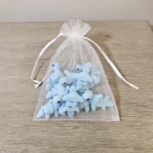 May include: A collection of light blue, miniature, stylized dinosaur figurines inside a sheer white drawstring bag. The bag is tied with a white ribbon and sits on a wooden surface.