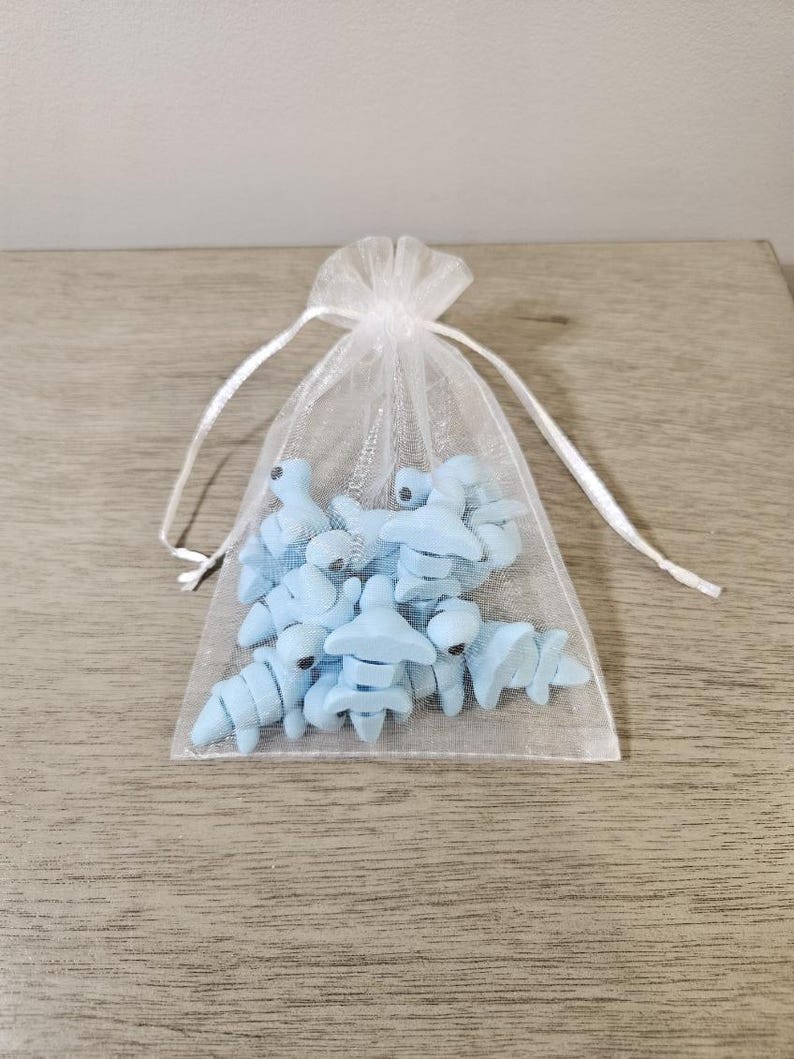 May include: A collection of light blue, miniature, stylized dinosaur figurines inside a sheer white drawstring bag. The bag is tied with a white ribbon and sits on a wooden surface.