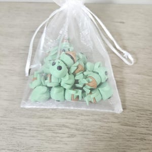 Puede incluir: Una colecci&oacute;n de peque&ntilde;as figuras de color verde claro que se asemejan a un personaje popular, dentro de una bolsa blanca transparente con cord&oacute;n. Las figuras tienen ojos negros y detalles marrones. La bolsa est&aacute; sobre una superficie de madera gris claro.