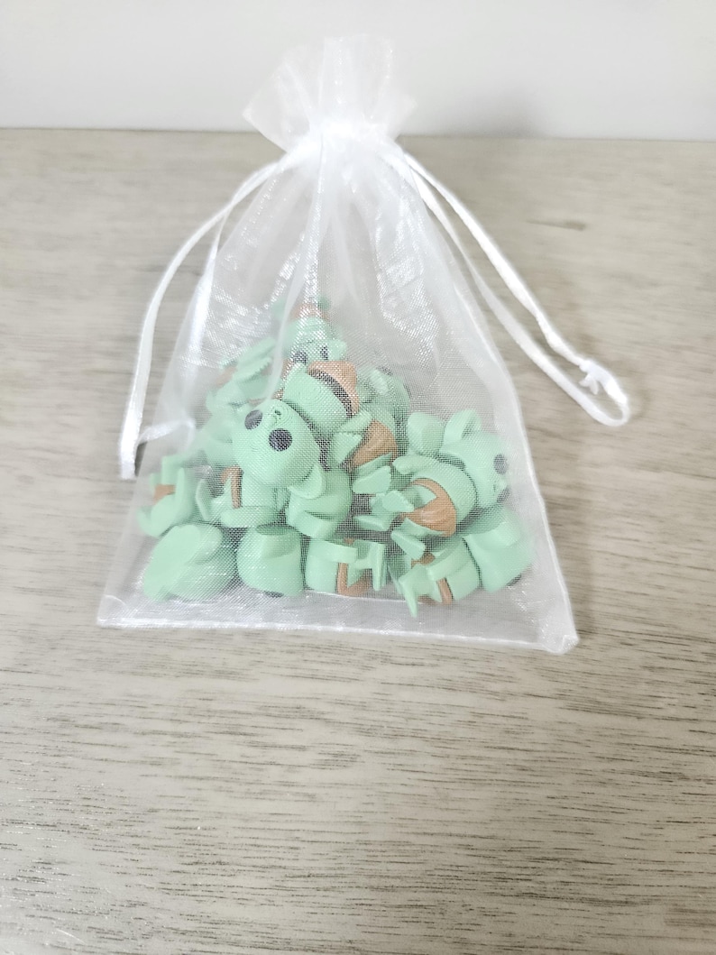 Puede incluir: Una colecci&oacute;n de peque&ntilde;as figuras de color verde claro que se asemejan a un personaje popular, dentro de una bolsa blanca transparente con cord&oacute;n. Las figuras tienen ojos negros y detalles marrones. La bolsa est&aacute; sobre una superficie de madera gris claro.