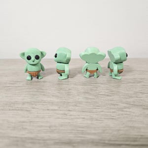 Puede incluir: Cuatro peque&ntilde;as figuras verdes de un personaje con orejas grandes y ojos grandes, que lleva un pa&ntilde;al marr&oacute;n. Las figuras est&aacute;n en diferentes poses, mostrando al personaje desde varios &aacute;ngulos. Las figuras est&aacute;n sobre una superficie marr&oacute;n claro.