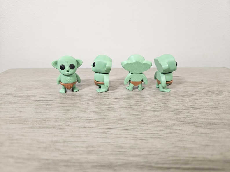 Puede incluir: Cuatro peque&ntilde;as figuras verdes de un personaje con orejas grandes y ojos grandes, que lleva un pa&ntilde;al marr&oacute;n. Las figuras est&aacute;n en diferentes poses, mostrando al personaje desde varios &aacute;ngulos. Las figuras est&aacute;n sobre una superficie marr&oacute;n claro.