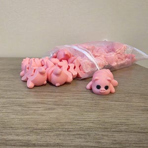 May include: A collection of pink pig-shaped craft supplies. Several pigs are scattered on a wooden surface, with a larger quantity inside a clear plastic bag. Each pig has small black eyes.