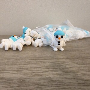 May include: A collection of small, white snowman figurines with blue hats and black button eyes. One snowman stands upright, while others are scattered on a wooden surface. A mesh bag filled with more snowmen is in the background.