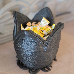 May include: A black, textured bowl filled with individually wrapped yellow and white candies. The bowl has a unique, organic shape with a textured exterior and a base with curled tendrils.