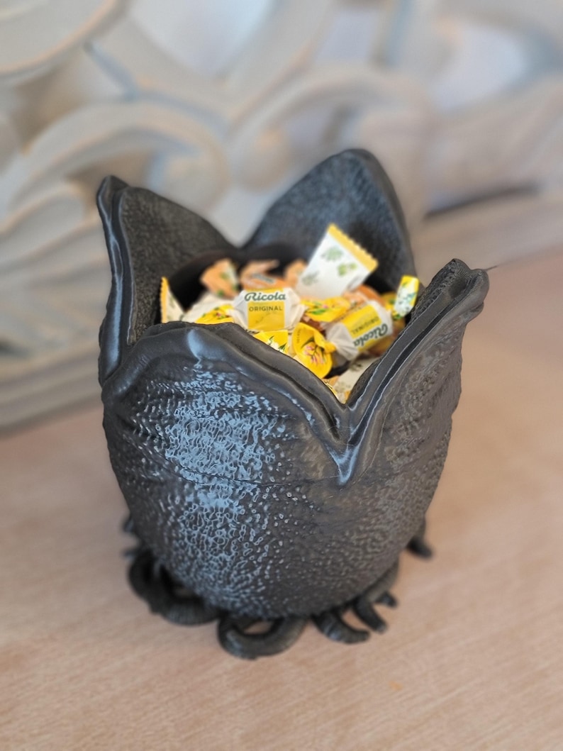 May include: A black, textured bowl filled with individually wrapped yellow and white candies. The bowl has a unique, organic shape with a textured exterior and a base with curled tendrils.