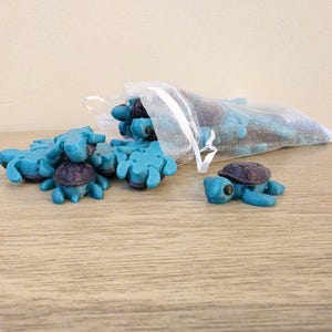 May include: A collection of small, teal turtle figurines with dark purple shells. Some turtles are scattered on a wooden surface, while others are in a clear, drawstring bag. The figurines are detailed and appear to be handmade.