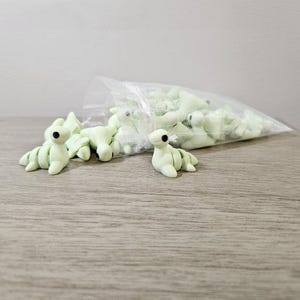 May include: A collection of light green, handmade clay dinosaur figurines with black eyes. Some are scattered on a wooden surface, while others are in a clear plastic bag. The figurines have a whimsical, cartoonish design.