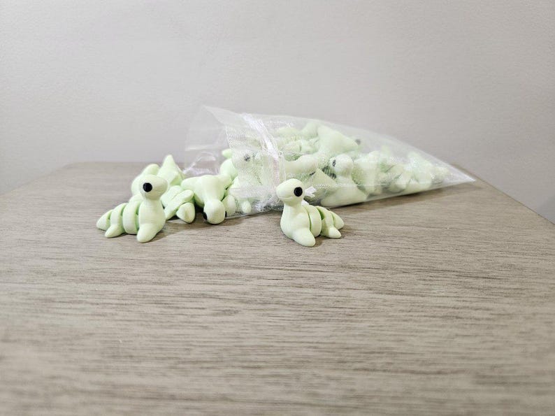 May include: A collection of light green, handmade clay dinosaur figurines with black eyes. Some are scattered on a wooden surface, while others are in a clear plastic bag. The figurines have a whimsical, cartoonish design.