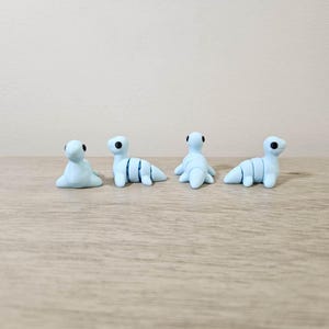 May include: Four small, light blue clay sculptures of a sea creature, each with black eyes. Two are in a seated position, and two are in a crawling position. The sculptures are on a light brown wooden surface.