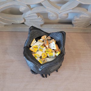 May include: A black, textured, decorative bowl filled with individually wrapped Ricola cough drops. The bowl has a unique, stylized shape. The background features a light-colored surface and ornate architectural details.