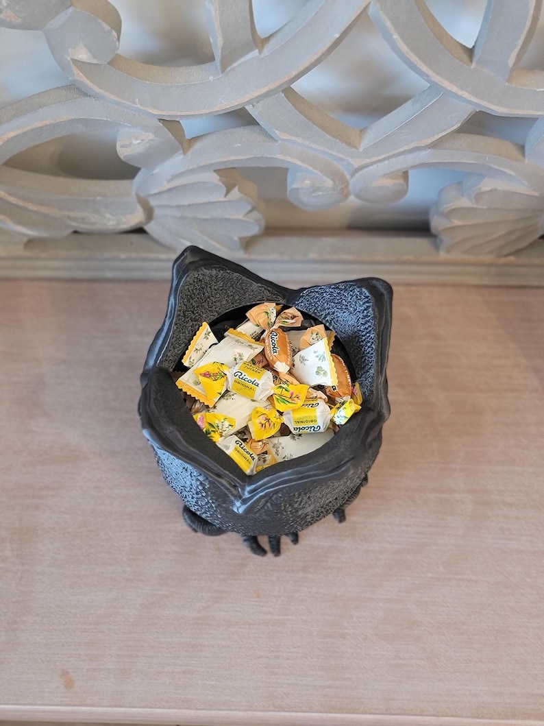 May include: A black, textured, decorative bowl filled with individually wrapped Ricola cough drops. The bowl has a unique, stylized shape. The background features a light-colored surface and ornate architectural details.