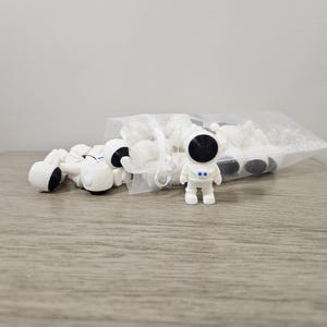 May include: A collection of small, white astronaut figurines with black visors and accents, some scattered and others in a clear plastic bag. The figurines are on a wooden surface.