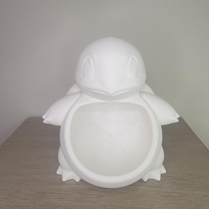 May include: A white ceramic figurine of a cartoon turtle character, featuring a round body, small arms, and a detailed face. The figurine is sitting on a wooden surface.