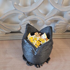 May include: A black, textured bat-shaped bowl filled with individually wrapped candies. The bowl has a wide opening and is resting on a wooden surface. The background features a decorative, light-colored wall element.