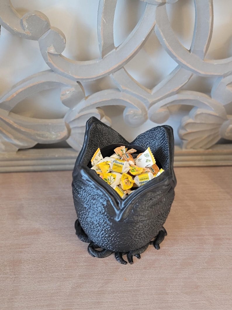 May include: A black, textured bat-shaped bowl filled with individually wrapped candies. The bowl has a wide opening and is resting on a wooden surface. The background features a decorative, light-colored wall element.