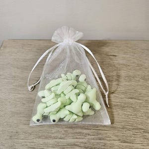 May include: A sheer white organza bag filled with light green frog-shaped candies. The candies have small black details and are presented on a wooden surface.