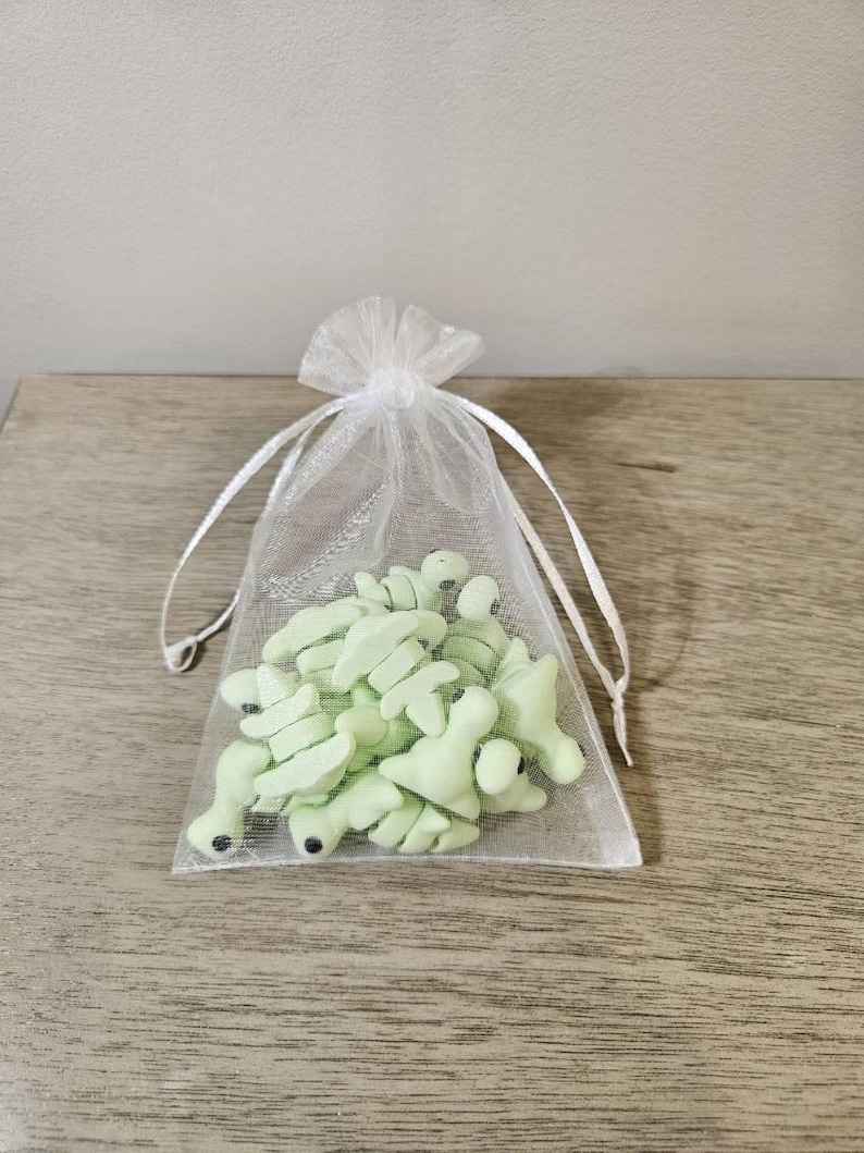 May include: A sheer white organza bag filled with light green frog-shaped candies. The candies have small black details and are presented on a wooden surface.