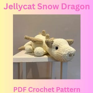 May include: A crocheted white and tan dragon toy with the text "Jellycat Snow Dragon" and "PDF Crochet Pattern". The dragon has tan wings, horns, and spikes along its back. The dragon is lying on a white surface.