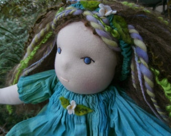 May include: A handmade doll with long brown hair, wearing a teal blue dress with a flower embellishment. The doll has blue eyes and a light complexion.