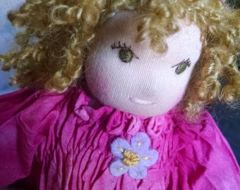 May include: A handmade doll with curly blonde hair, wearing a pink dress with a purple flower embroidered on the front. The doll has a sweet, embroidered face with closed eyes.
