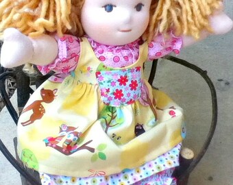 May include: A handmade doll with blonde yarn hair wearing a yellow dress with a pink and floral print. The dress has a layered skirt with a pink and white polka dot pattern.